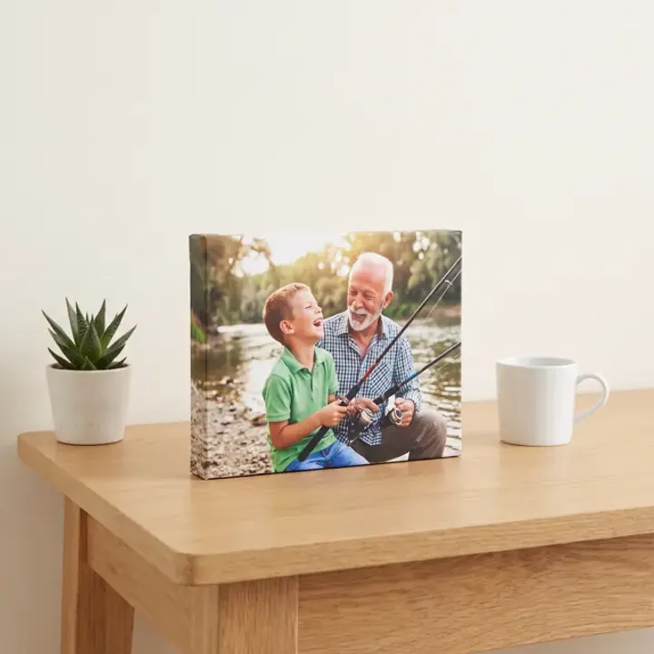 A desktop canvas of a grandad and grandson fishing on a small oak table beside a mug and a plant