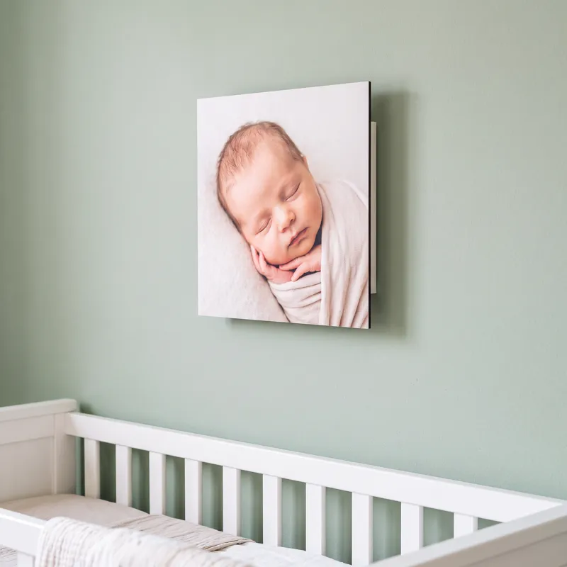 A single Floto photo tile of a sleeping newborn on a sage green nursery wall above a white cot
