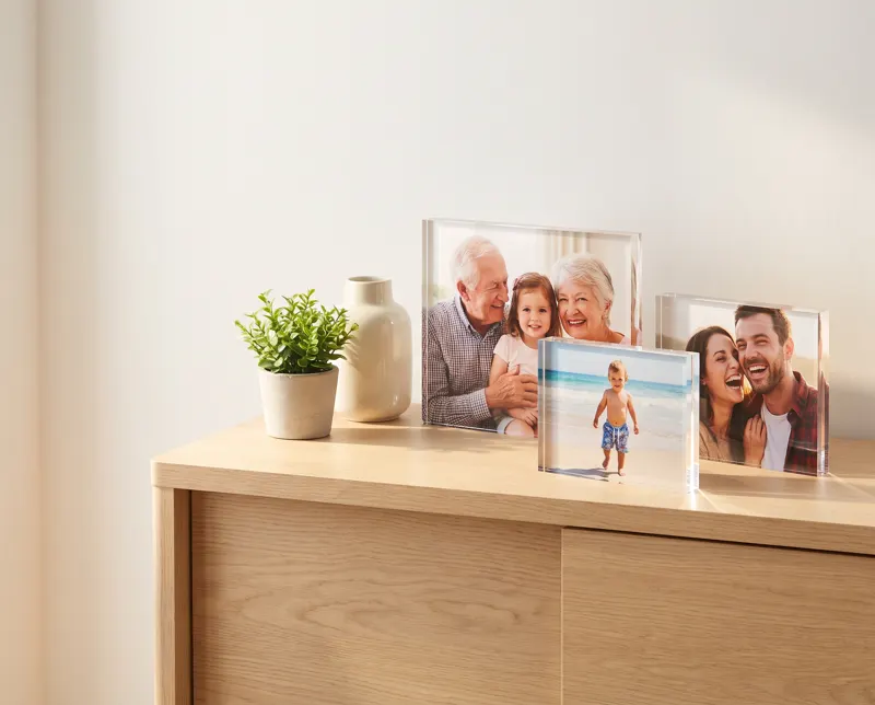 Three acrylic photo blocks on a pale oak sideboard: grandparents with a grandchild, a toddler on a beach and a laughing couple, each printed edge to edge behind clear acrylic