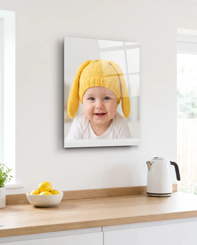 A high gloss metal print of a smiling baby in a yellow knitted bunny hat, on a white kitchen wall above a kettle