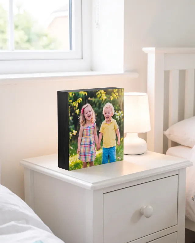 A black-edged wooden photo block of two laughing children in a sunny garden, on a white bedside table beside a lamp