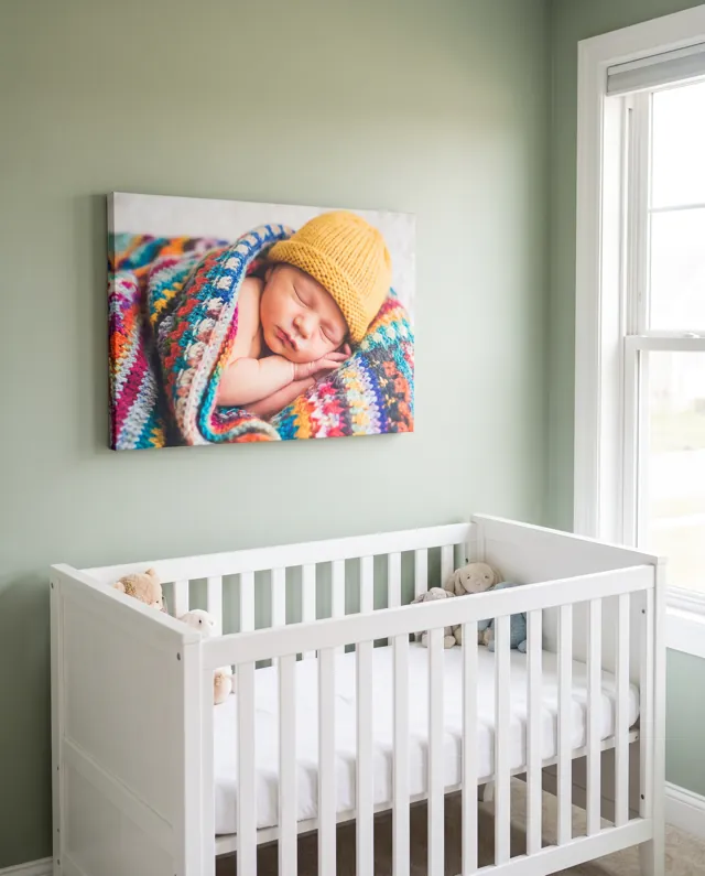 A canvas print of a sleeping newborn in a yellow knitted hat and a colourful blanket, above a white cot in a sage green nursery