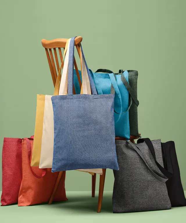 Recycled cotton tote bags in natural, black and soft marl colours, hung over a wooden chair