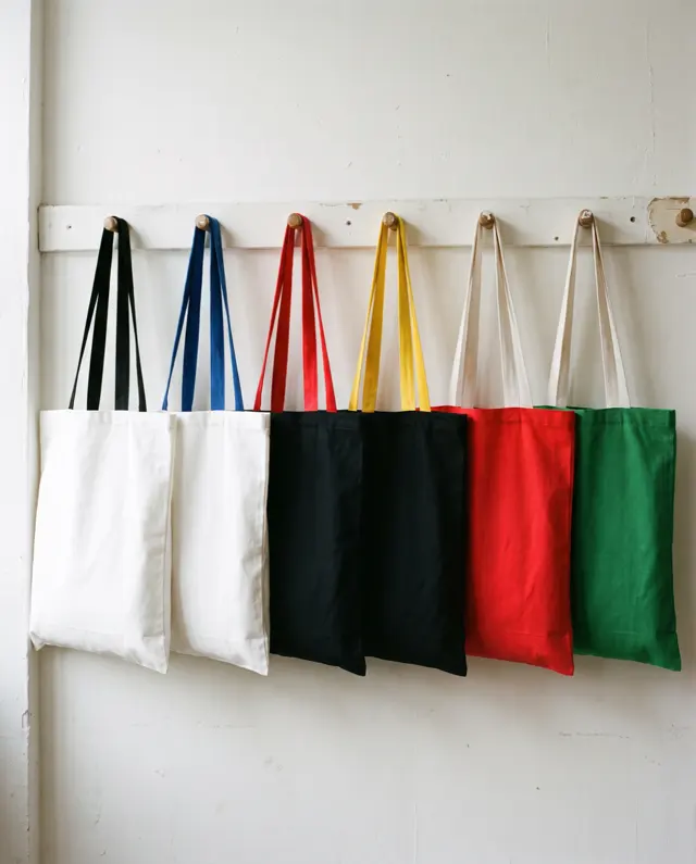 Six two tone varsity tote bags with contrast handles hanging from pegs on a white wall