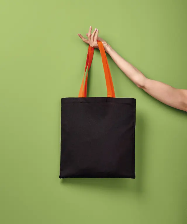 A black varsity tote bag with orange contrast handles, held up by its handles against a green wall